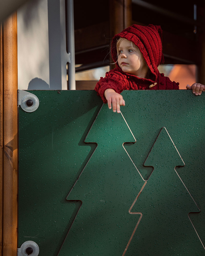 Small girl in red hoodie, overlooks from a playground unit which is forest themed. The panels have trees etched on the sides in HPL.